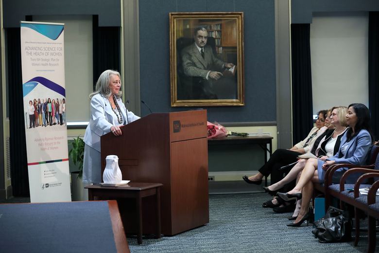 A woman stands behind a podium on the left side of the image. On the right, the first row of the audience is seen sitting in chairs.
